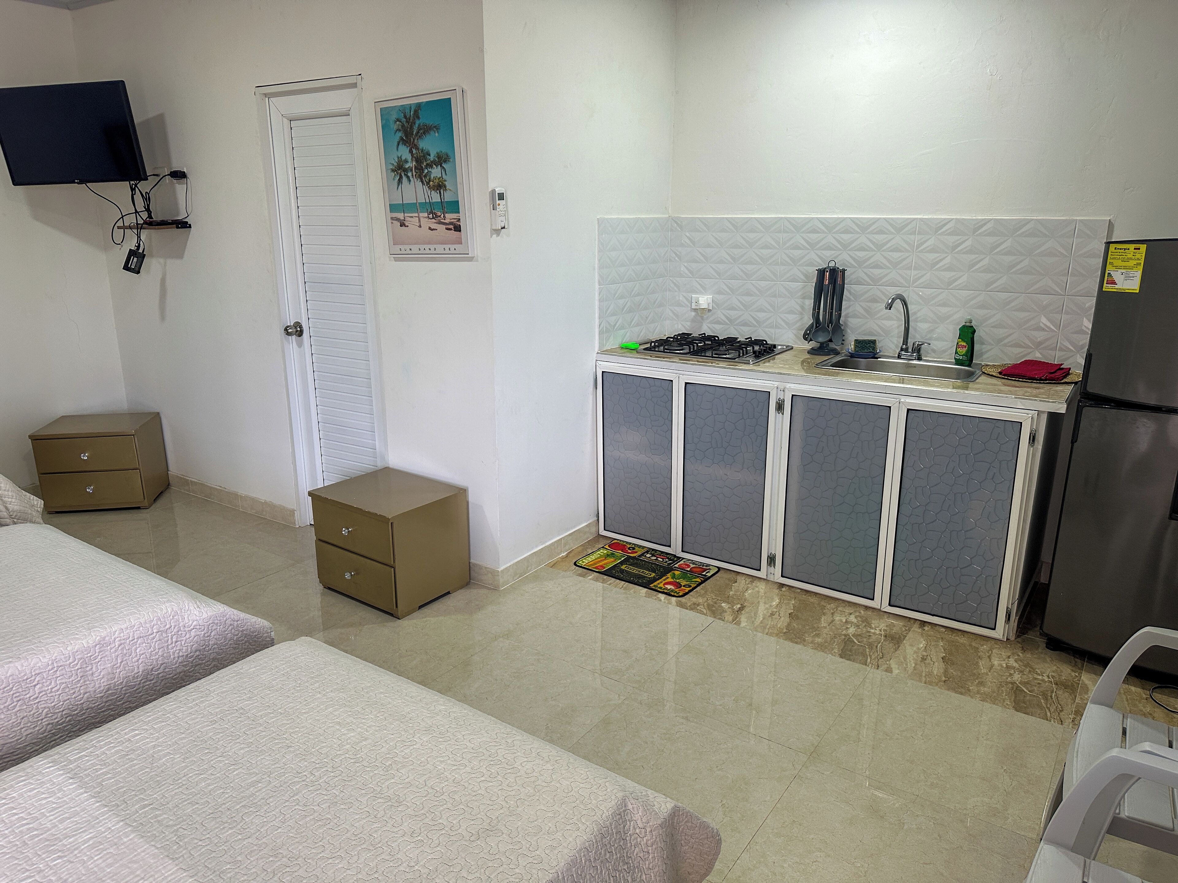 A compact hotel room with a kitchenette, a bed, a small bedside table, a nightstand, and a TV mounted on the wall.