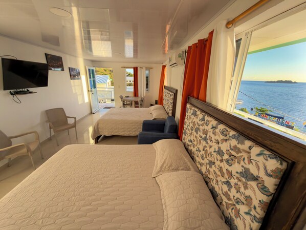 Ein Hotelzimmer mit zwei Betten, einem Fernseher, einem Balkon mit Meerblick und einem Essbereich.