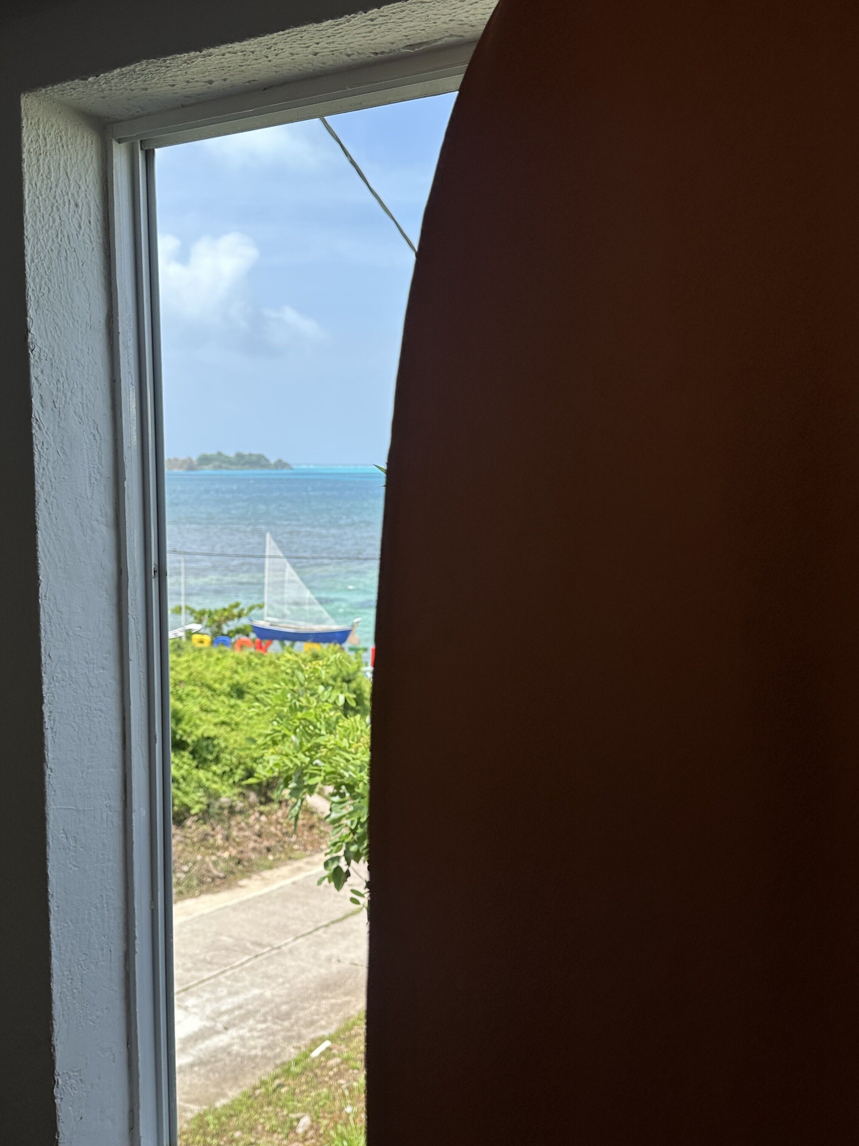 Blick durch ein Fenster auf das Meer, ein Boot und etwas Grün.