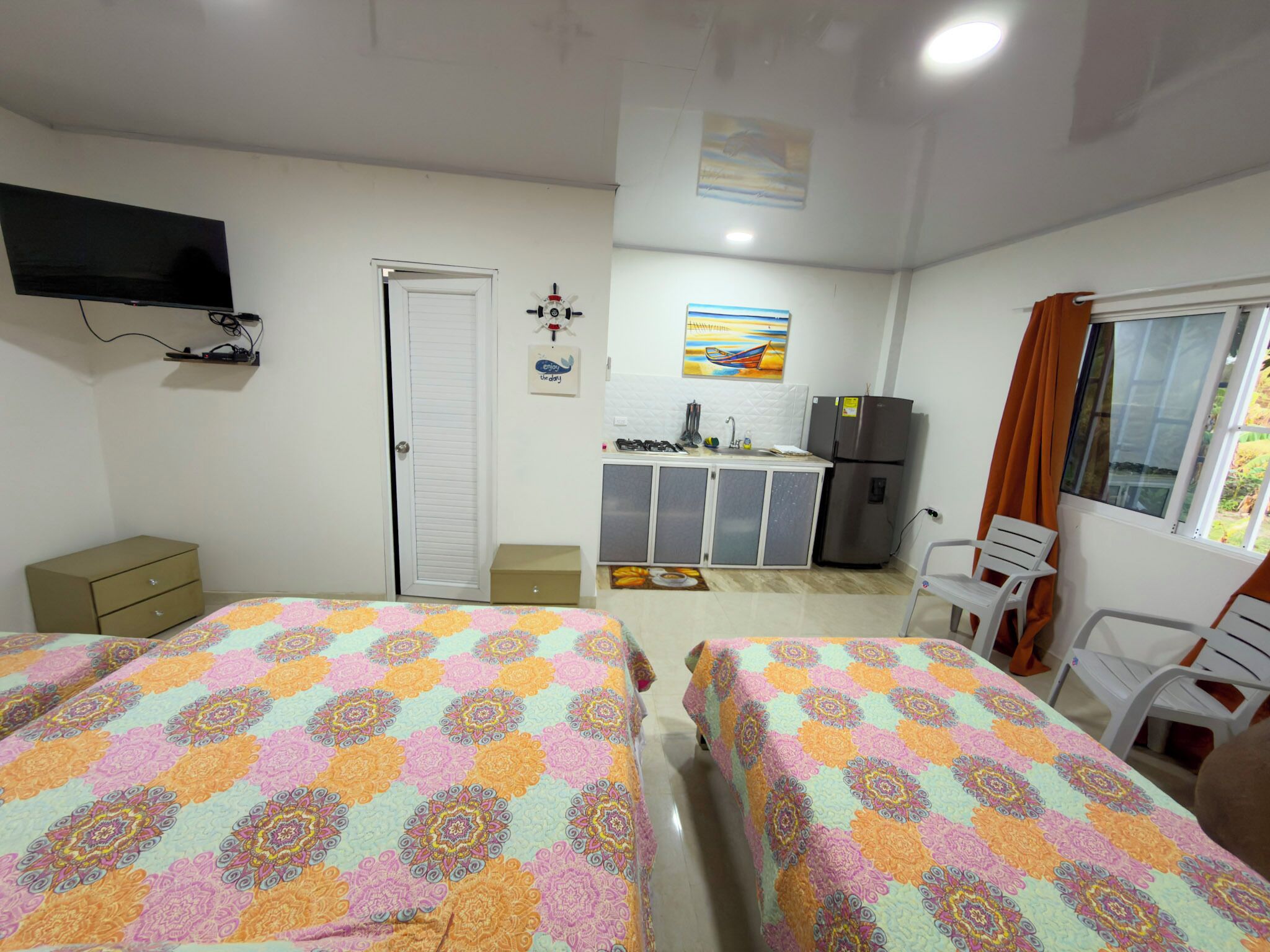 A room with two beds, a TV, a kitchenette, a window with curtains, and a painting on the wall.