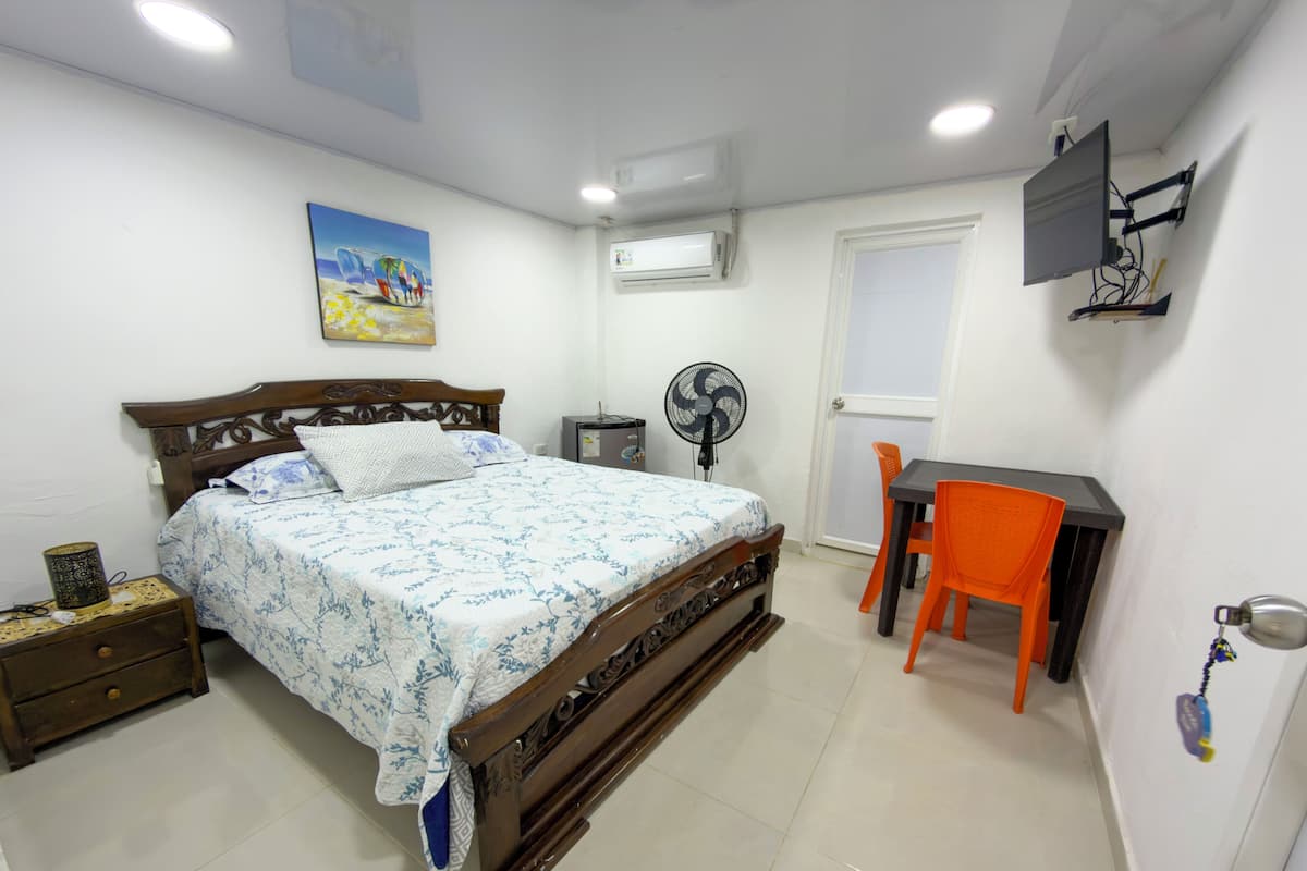 Un dormitorio con cama, mesita de noche, ventilador, escritorio con silla y televisor instalado en la pared.
