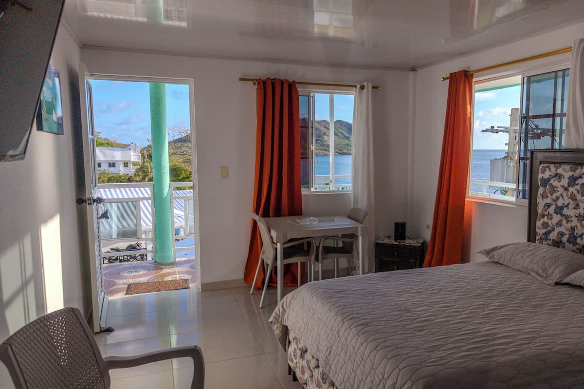 Un dormitorio con cama, escritorio y silla, un balcón con vistas al mar y una ventana con cortinas.