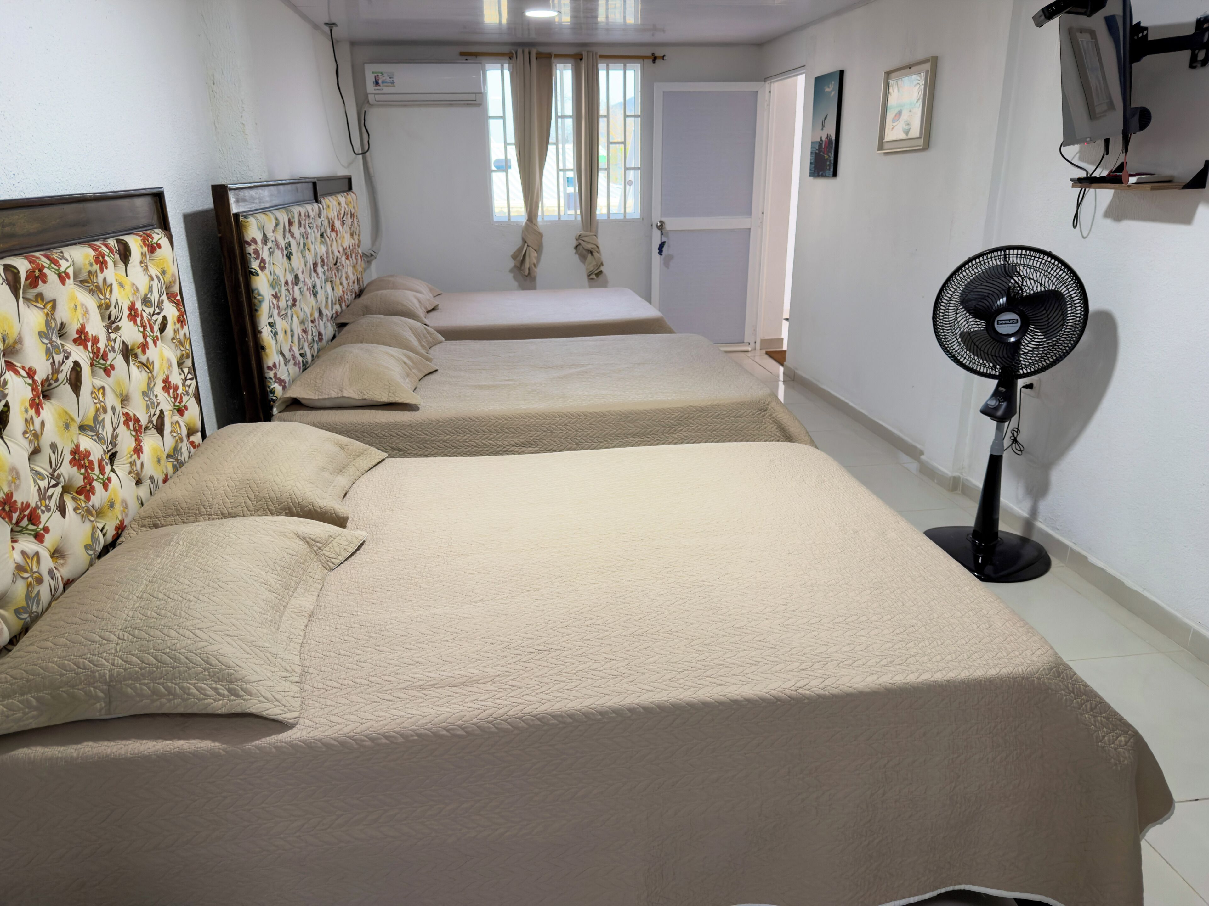 Habitación de hotel con tres camas, un ventilador de pie y una ventana con cortinas.