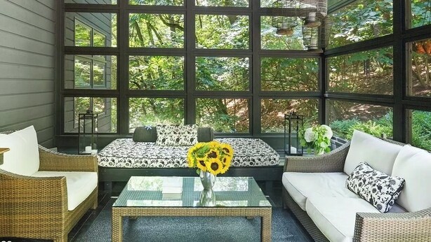 A covered porch with wicker furniture, a glass coffee table, and a view of a forest through large windows.