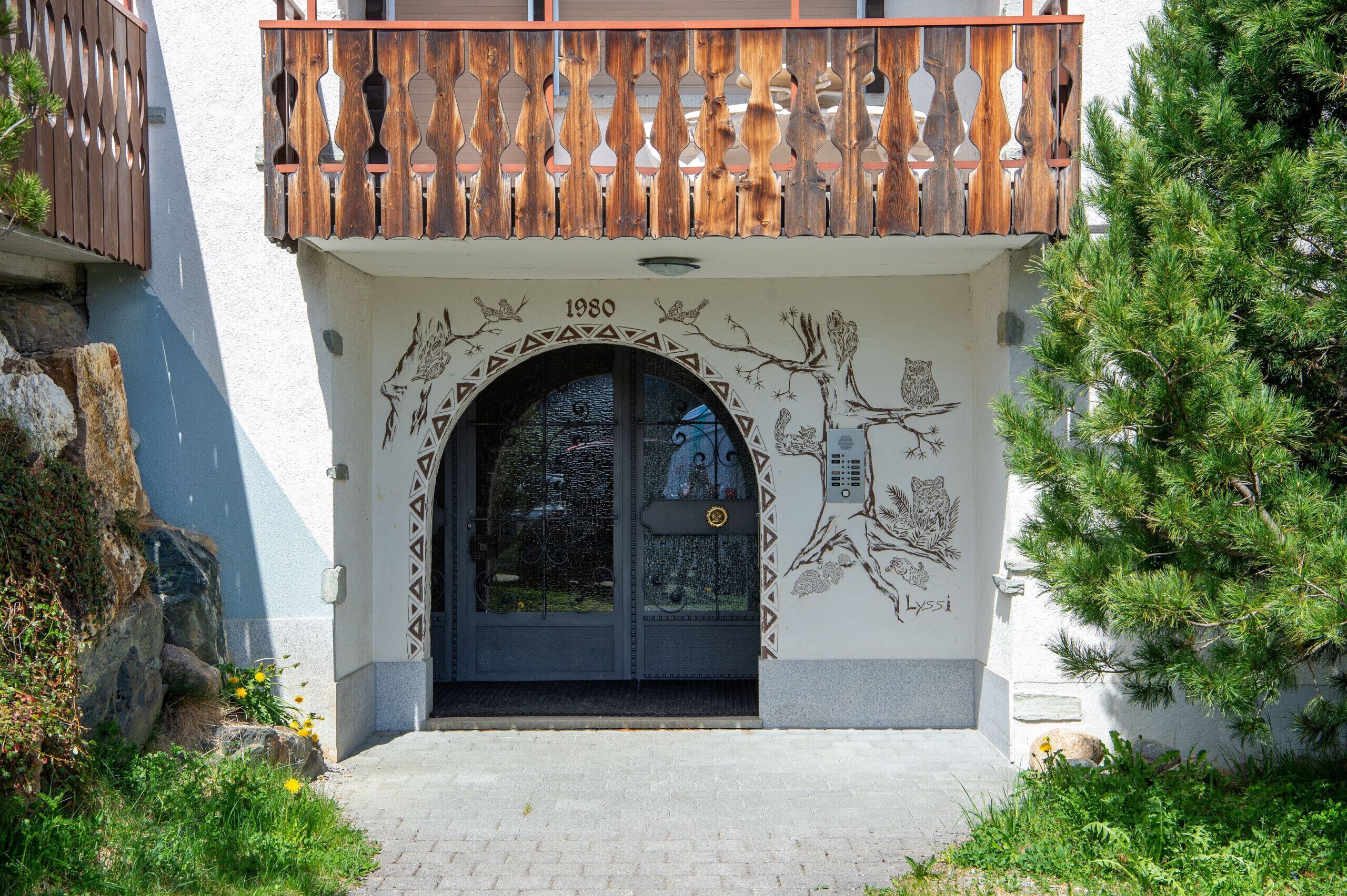 L’entrée d’un immeuble, avec un balcon en bois, une porte dotée d’un panneau de verre et une fresque représentant des arbres et des hiboux.