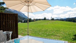 A table with a glass top and a beige umbrella, overlooking a grassy field and mountains.