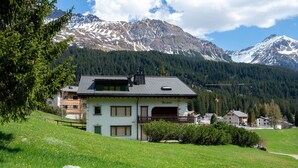 A two-story building with a balcony, surrounded by a green landscape and mountains in the background.