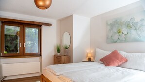 A bedroom with a large bed, a window with a view of greenery, a bedside table with a lamp, and a wall-mounted mirror.