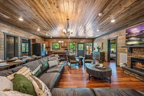 A spacious living room with a large sofa, a coffee table, and a fireplace. There is a kitchen area with a refrigerator and cabinets visible through the open door. - Luxe 8BR w/Pool,Outdoor Play Area, Huge Sports Bar (Sevierville)