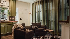 Un salón acogedor con dos sillones oscuros, una mesita redonda y una alfombra estampada. Se observa una zona de cocina con armarios verdes y una planta colgante.
