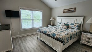 A bedroom with a bed, two bedside tables, a TV mounted on the wall, and a window with blinds.