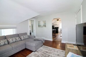 A modern living room with a sectional sofa, a flat-screen TV, and a dining area in the background. - Recovery Loft 1-bedroom apartment in Mansfield, OH with WiFi, AC, Full Kitchen (Mansfield)