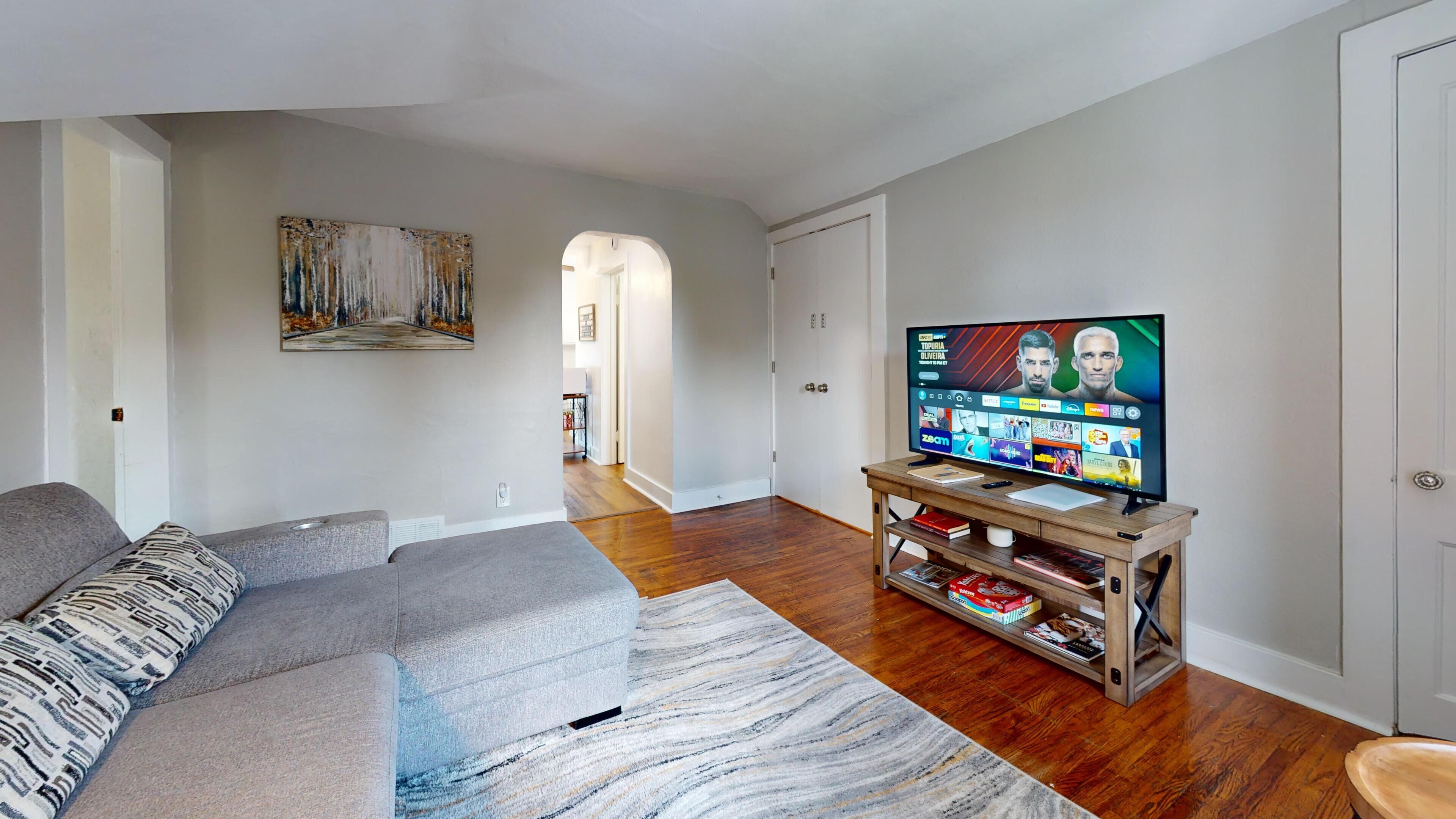 Un salón con un sofá gris, un mueble de televisión de madera y un televisor de pantalla plana grande que muestra un evento deportivo.