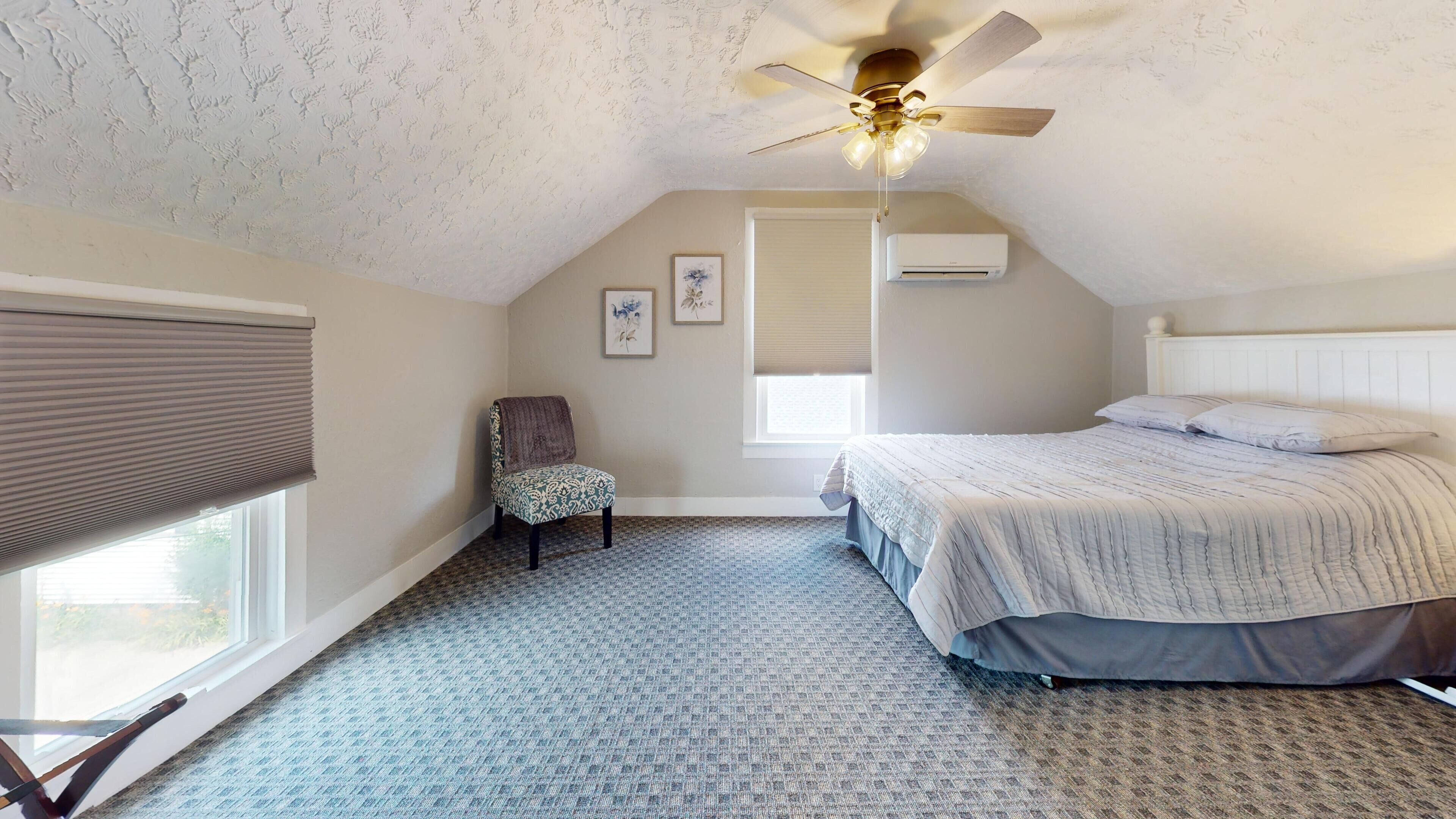 A bedroom with a sloped ceiling, a ceiling fan, a bed with a patterned bedspread, a chair, and a window with blinds.