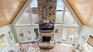 Una habitación con techos altos, chimenea de piedra, ventanas amplias y vista a un lago.
