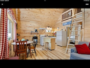A cozy cabin interior with a dining area, kitchen, and a loft bedroom with a ladder.