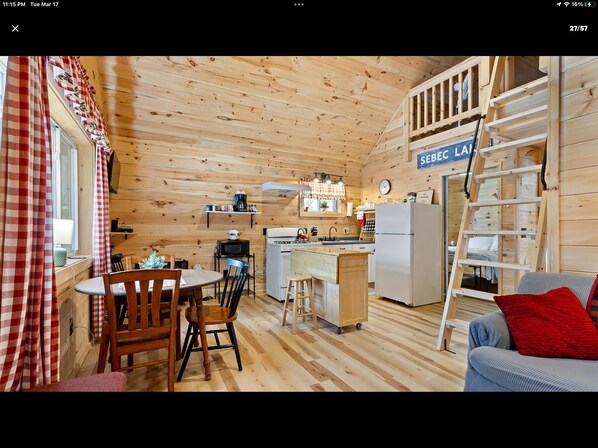 A cozy cabin interior with a dining area, kitchen, and a loft bedroom with a ladder.