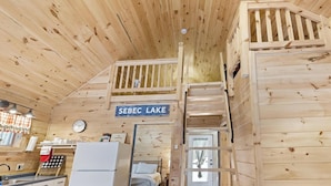 A wooden cabin interior with a loft bedroom, a kitchen area, and a sign indicating 'SEBEC LAKE'.