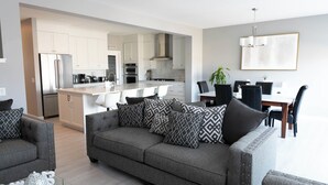 A modern living room with a grey sofa, patterned cushions, and a coffee table. A dining area with a table and chairs is visible in the background. - AC, Scenic Cochrane Family Home (Cochrane)