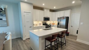 Una cocina moderna con armarios blancos, una isla central con taburetes de bar, un refrigerador de acero inoxidable y un microondas.