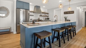 Een moderne keuken met een centraal eiland, barkrukken, roestvrijstalen apparatuur, hanglampen en een decoratief bord.