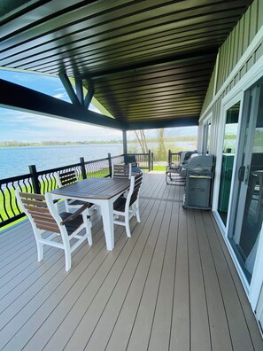 A covered outdoor deck with a table and chairs, a grill, and a view of a lake.
