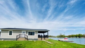 Een gelijkvloers huis met een overdekte veranda, een klein terras en een steiger die in een meer uitstelt.