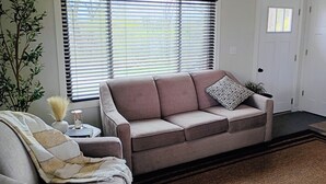 Een woonkamer met een beige bank, een kussen met patroon, een klein bijzettafeltje met een vaas, en een groot raam met jaloezieën.