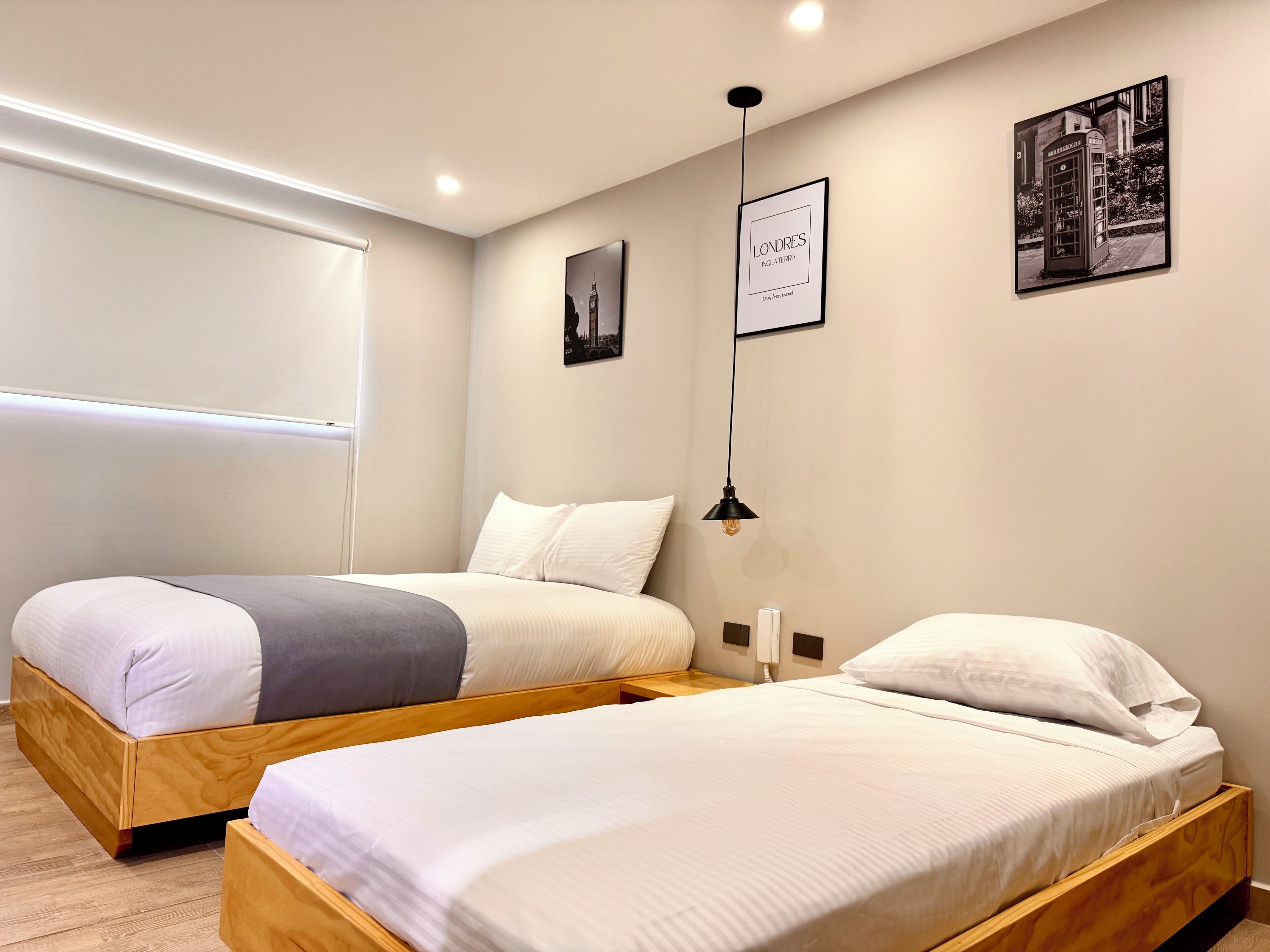 Habitación de hotel con dos camas de madera, ropa de cama blanca, una lámpara colgante negra y cuadros en las paredes.