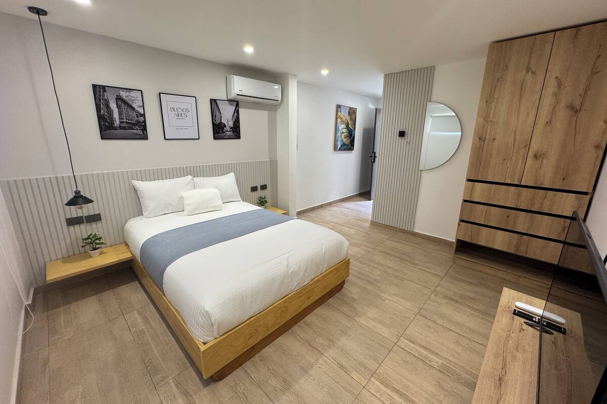 A modern bedroom with a wooden bed, a nightstand, a wall-mounted air conditioner, and a wardrobe with a mirror.