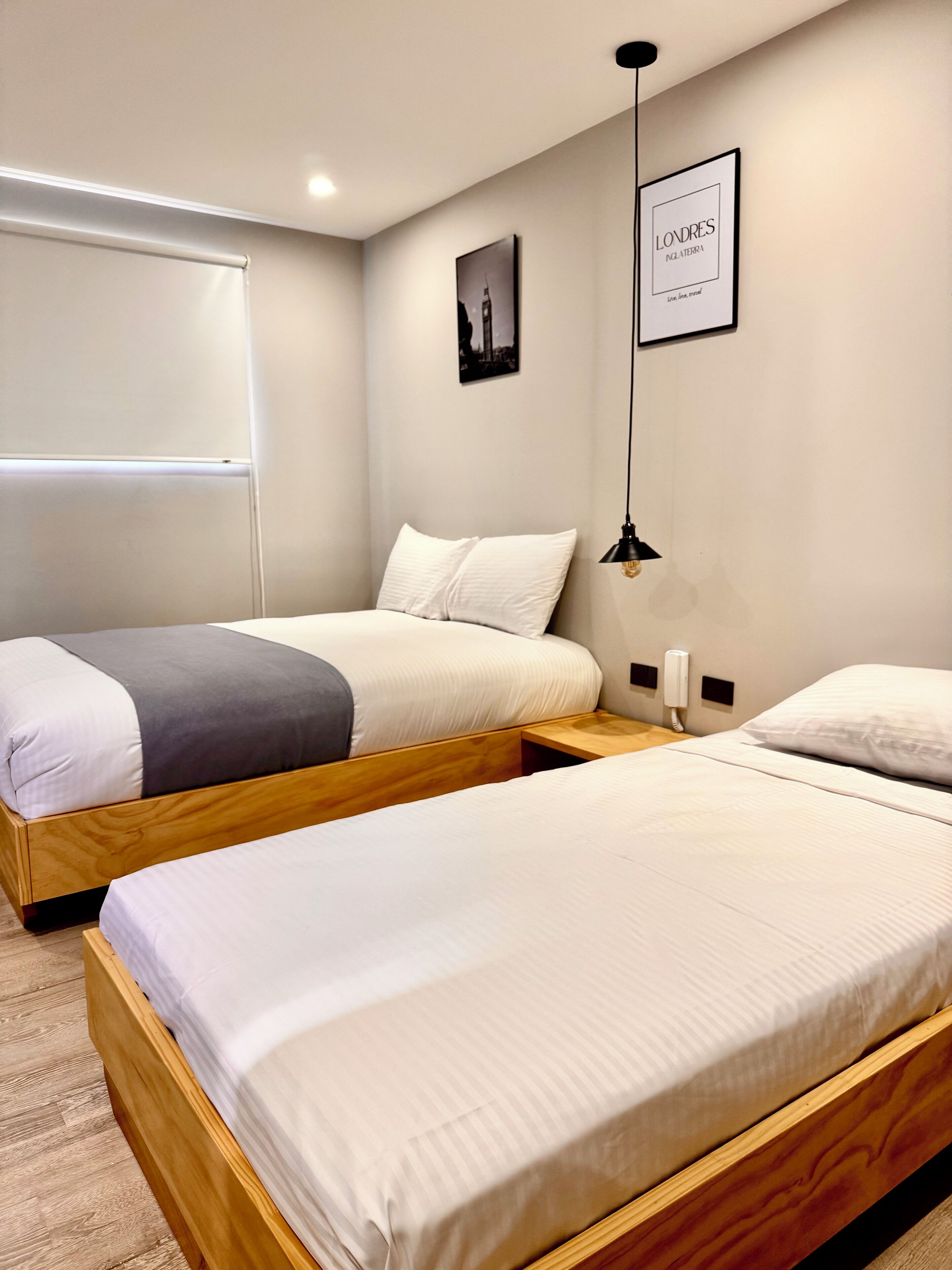 A hotel room with two wooden beds, a white wall, a framed picture, and a ceiling light.