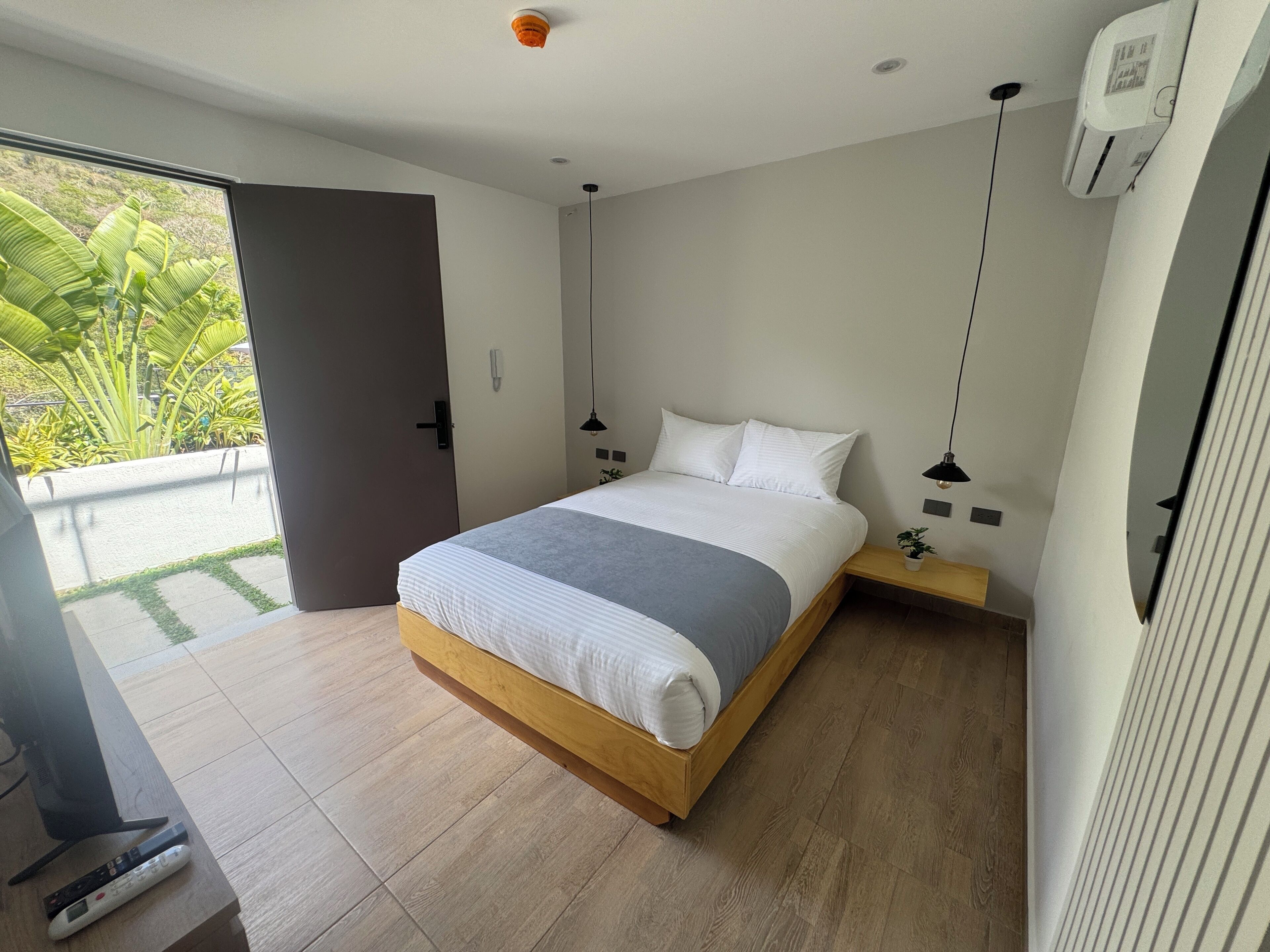 Un dormitorio moderno con cama de madera, una mesita de noche pequeña, aire acondicionado de pared y vistas a zonas verdes.