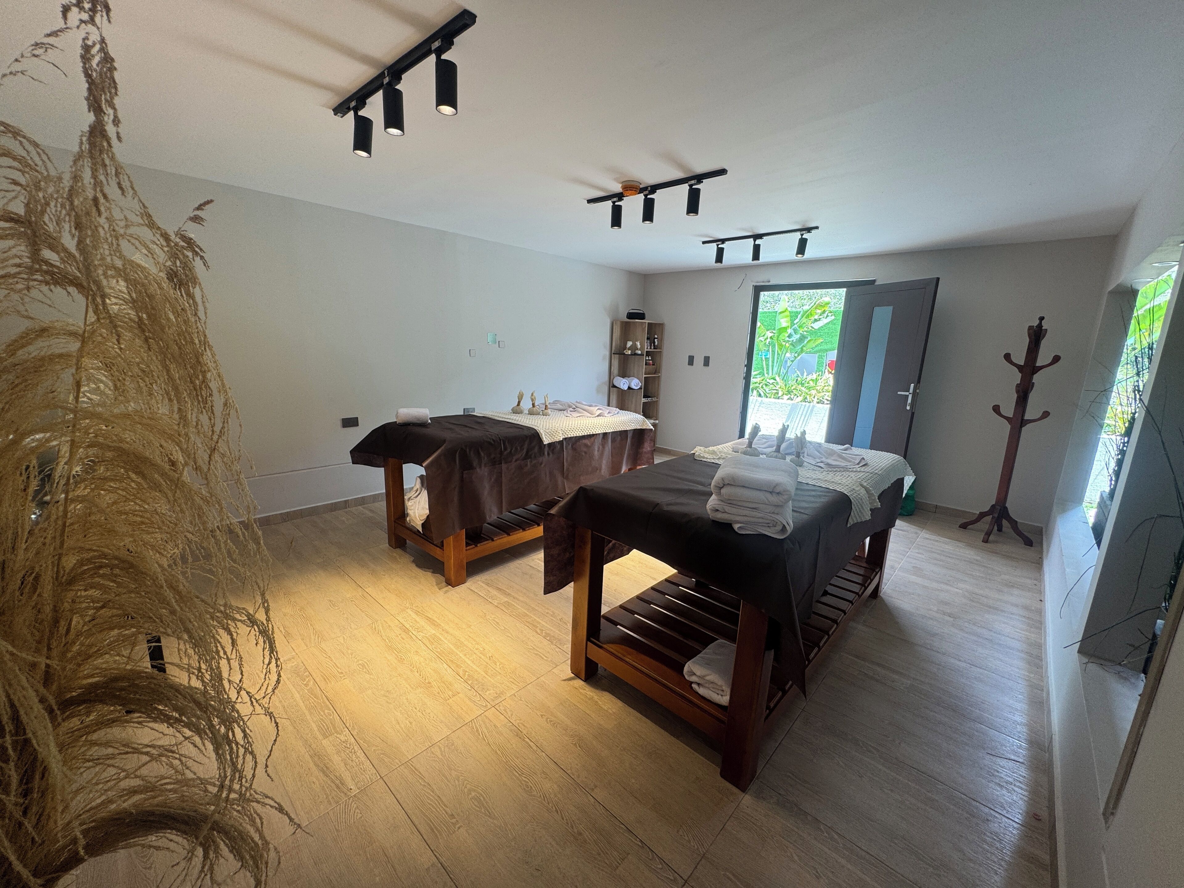 Une salle de spa équipée de deux tables de massage, d’un luminaire au plafond, d’un porte-manteau et offrant une vue sur la verdure environnante.