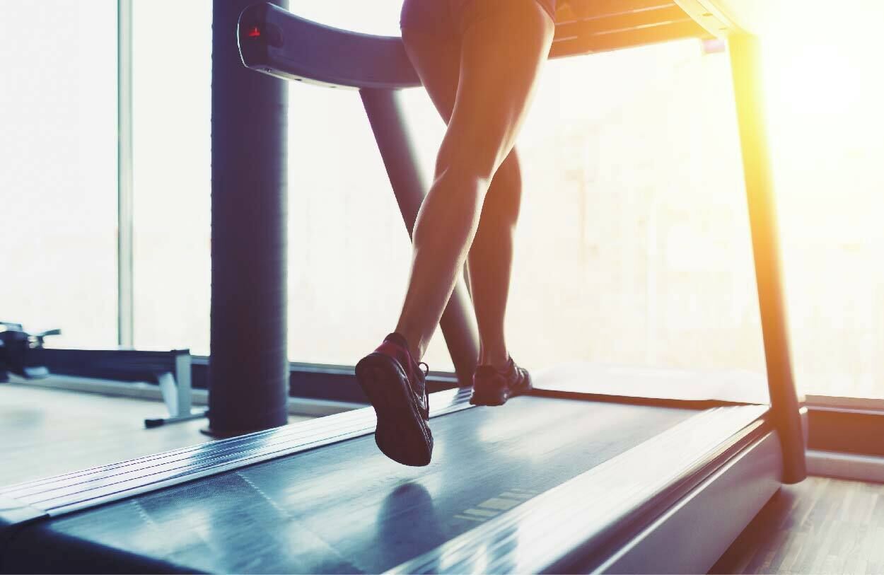 A person running on a treadmill in a gym with large windows.