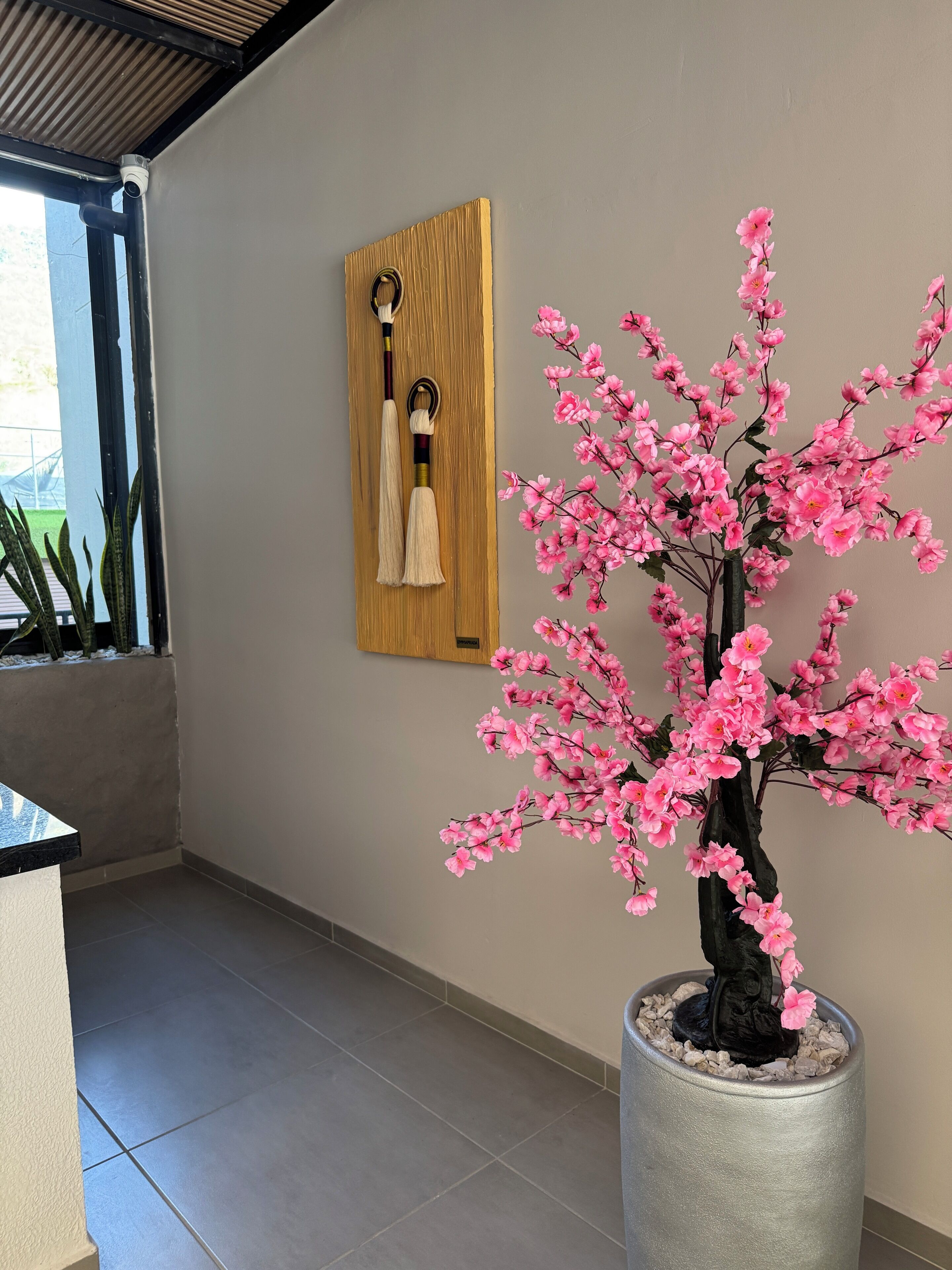A hallway with a potted tree with pink flowers, a wooden panel with tassels, and a security camera.