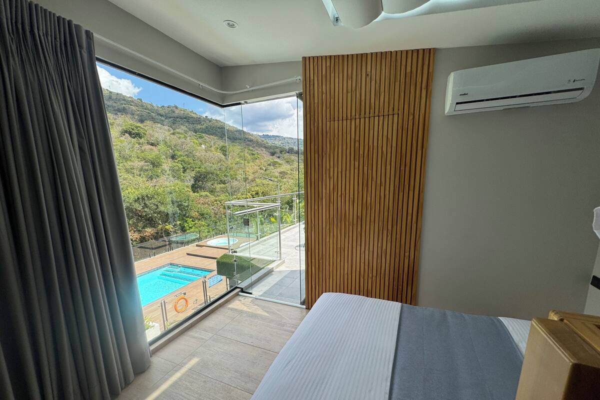 A hotel room with a bed, a view of a pool and mountains, and an air conditioning unit.