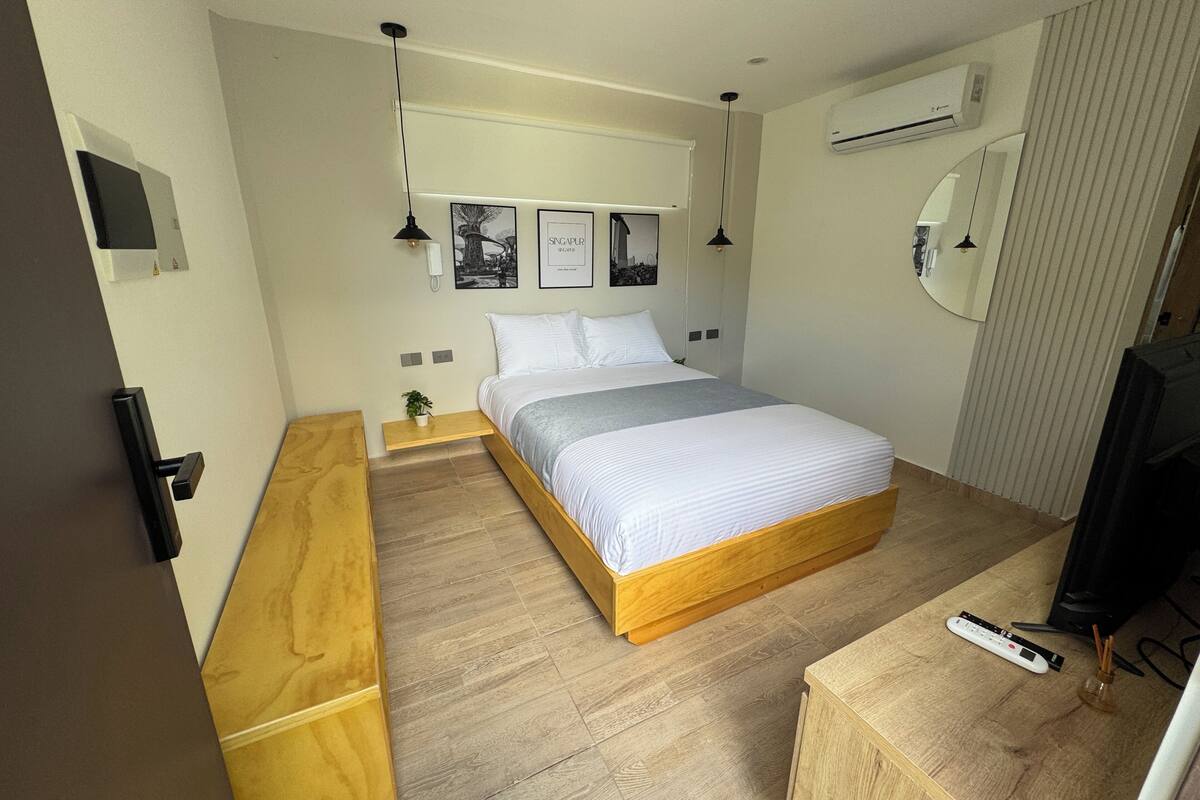 A modern hotel room with a wooden bed, bedside tables, a small plant, and an air conditioning unit.