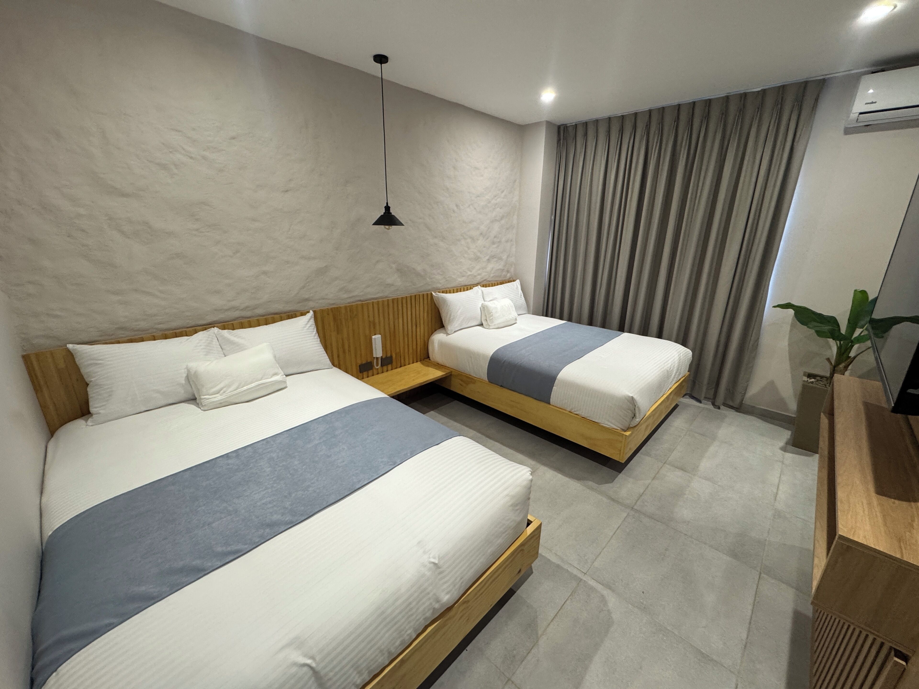 Une chambre d’hôtel avec deux lits, une tête de lit en bois, une table de chevet avec un téléphone, une plante et un luminaire moderne.