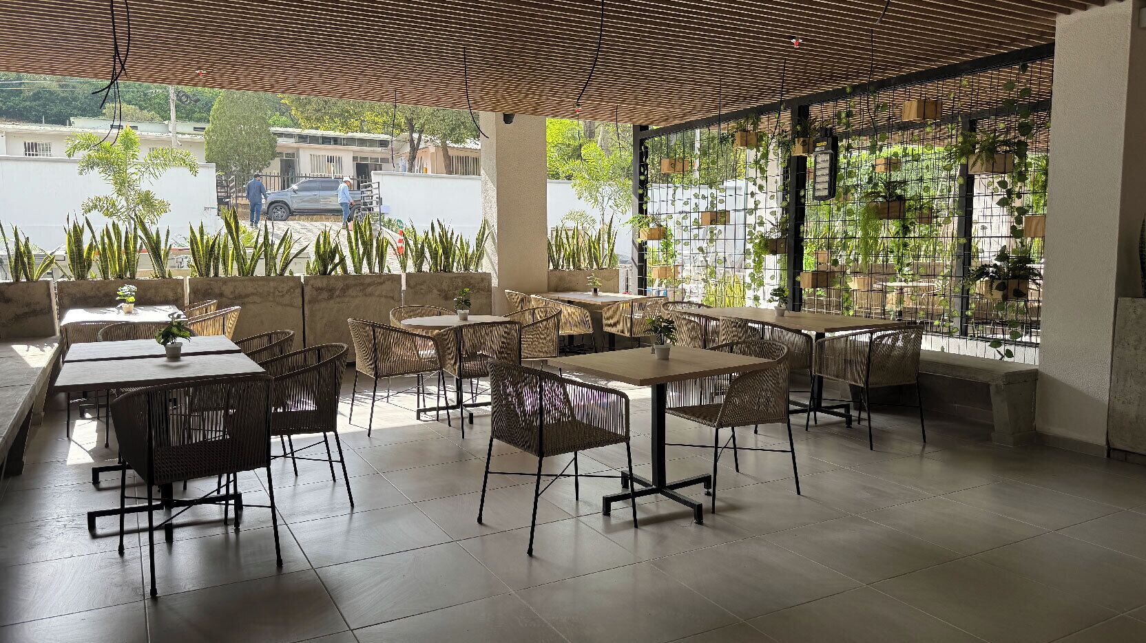 An outdoor patio with tables and chairs, surrounded by potted plants and a view of the street.