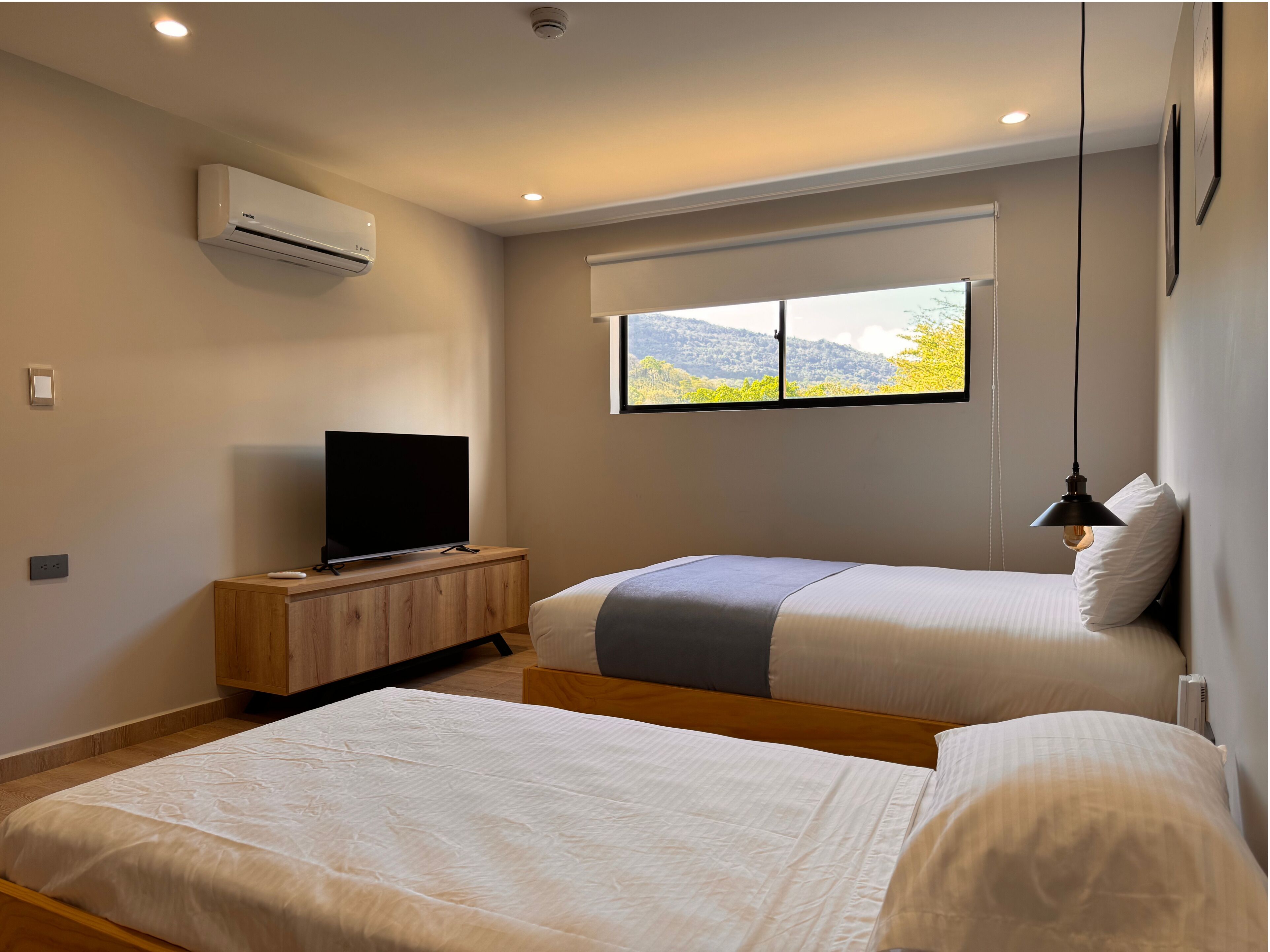 A modern hotel room with two beds, a television on a wooden cabinet, an air conditioning unit, and a window with a mountain view.