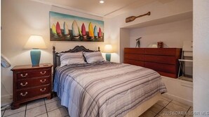 Un dormitorio con cama, dos mesitas de noche con lámparas, un aparador y un cuadro de veleros, además de un cartel de bienvenida.