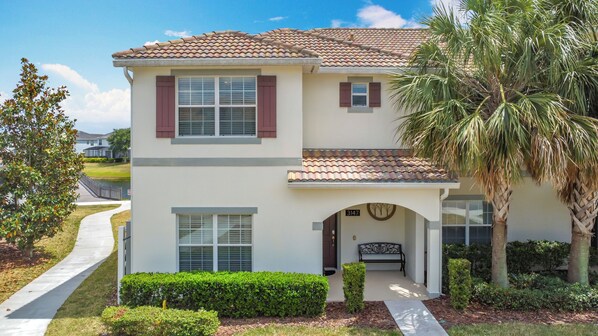 A two-story house with a tiled roof, a front porch with a bench, and a well-maintained lawn.