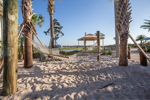 A hammock set up between palm trees on a sandy beach with a gazebo in the background.