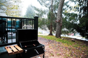 A barbecue grill with a metal grate and a backrest, placed outdoors with a view of trees and a bench in the background. - A-Frame: Fire Pit, Deck, grill & Lake Community (Tobyhanna)