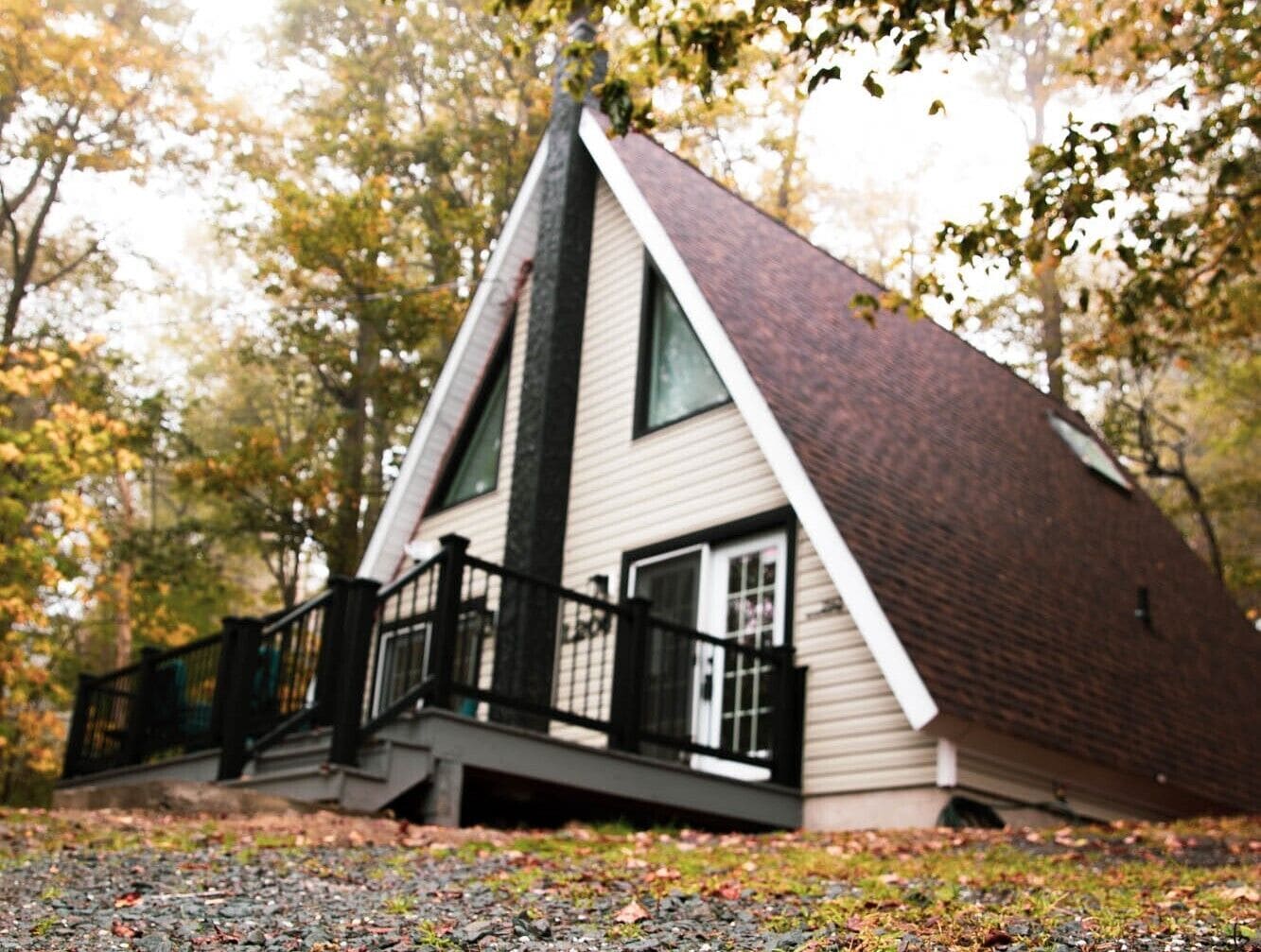 A cabin with a steeply pitched roof, a front porch with a black railing, and a large window on the side.