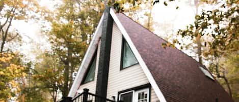 A cabin with a steeply pitched roof, a front porch with a black railing, and a large window on the side.
