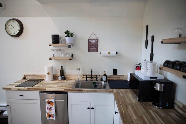 Una cocina compacta con encimeras de madera, un lavamanos de acero inoxidable y un lavavajillas. Hay estantes con cristalería y un cartel que dice 'KITCHEN'.