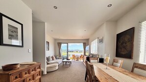 A spacious living area with a wooden dining table, a sofa set, and a view of the outdoors. - Charming Lakeview Golf Villa (Matarangi)