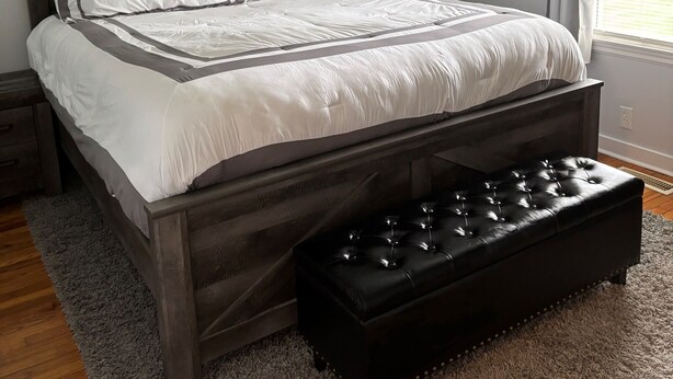 Un dormitorio con cama de madera, ventilador de techo, mesita de noche, un banco, una alfombra y una pared con palabras inspiradoras.