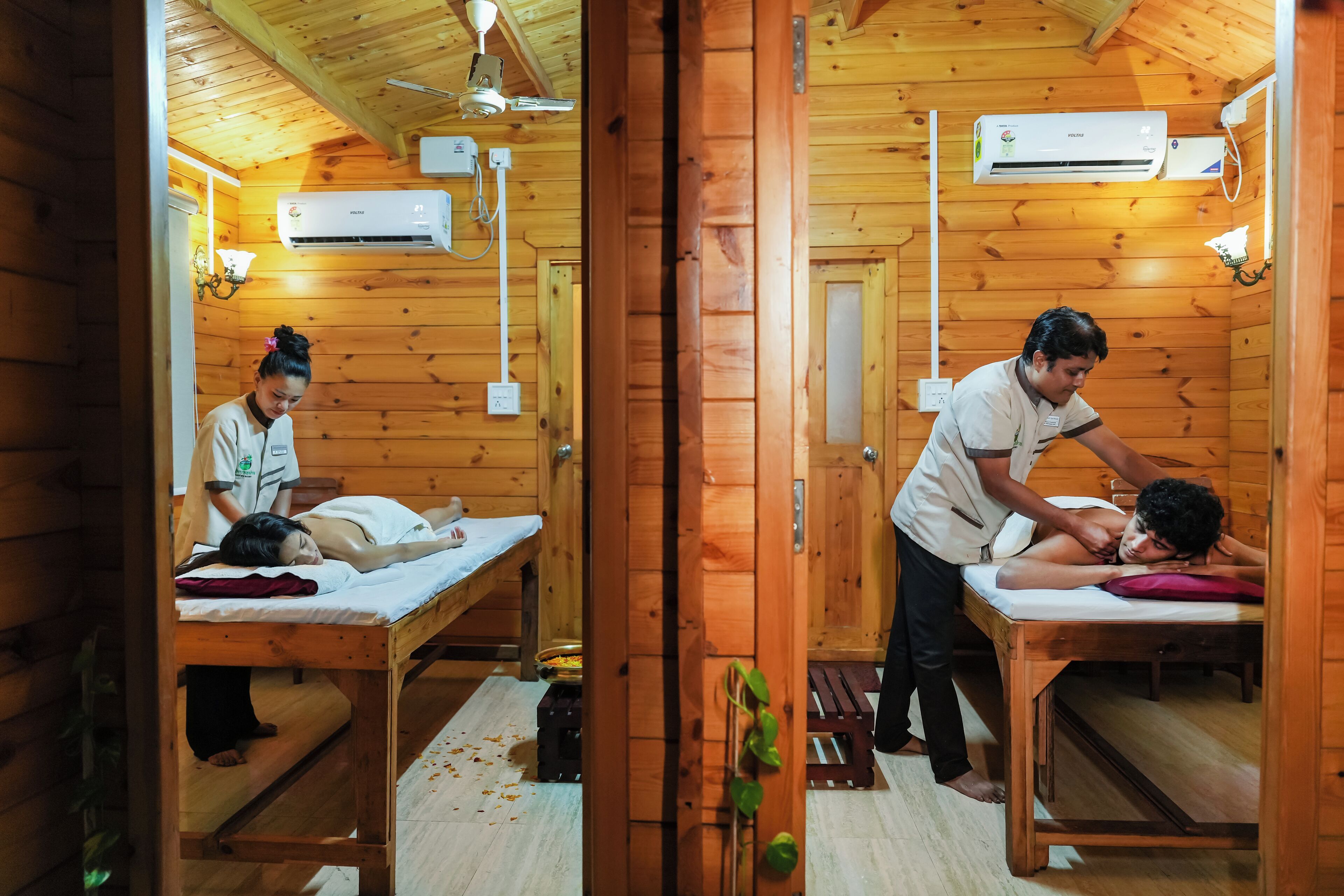 Una habitación de madera con dos mesas de masaje, cada una con una persona recibiendo un masaje. Se ven unidades de aire acondicionado en las paredes y un ventilador de techo.